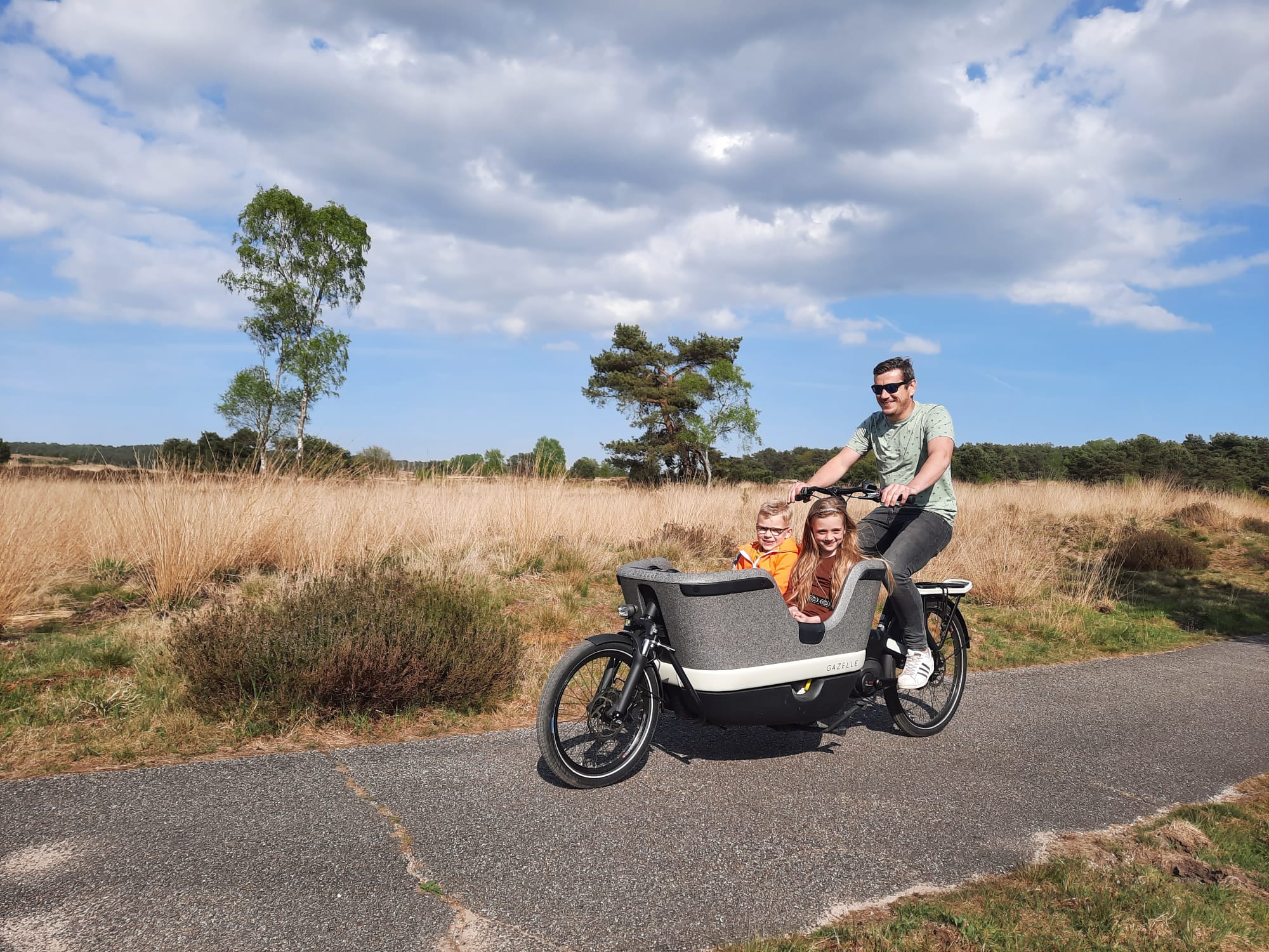 Man fietst met twee kinderen in een bakfiets op een pad door een natuurlijke omgeving.
