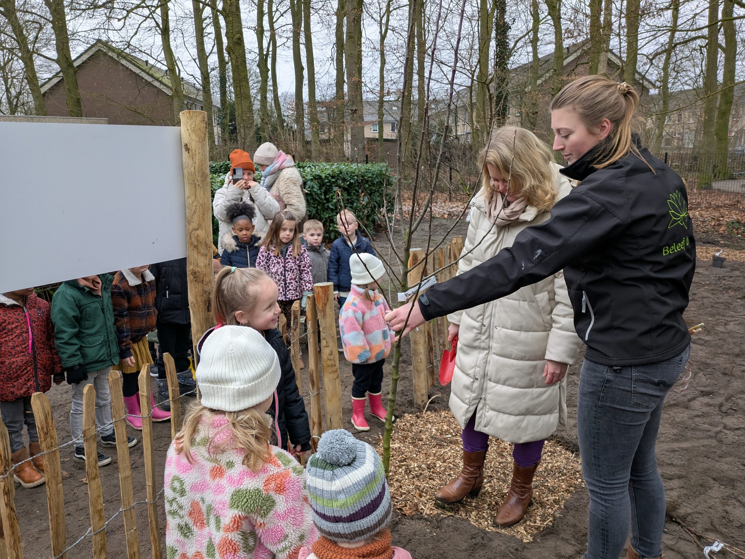 Kinderen planten een boom met begeleiding in een omheinde tuin in een bosrijke omgeving.