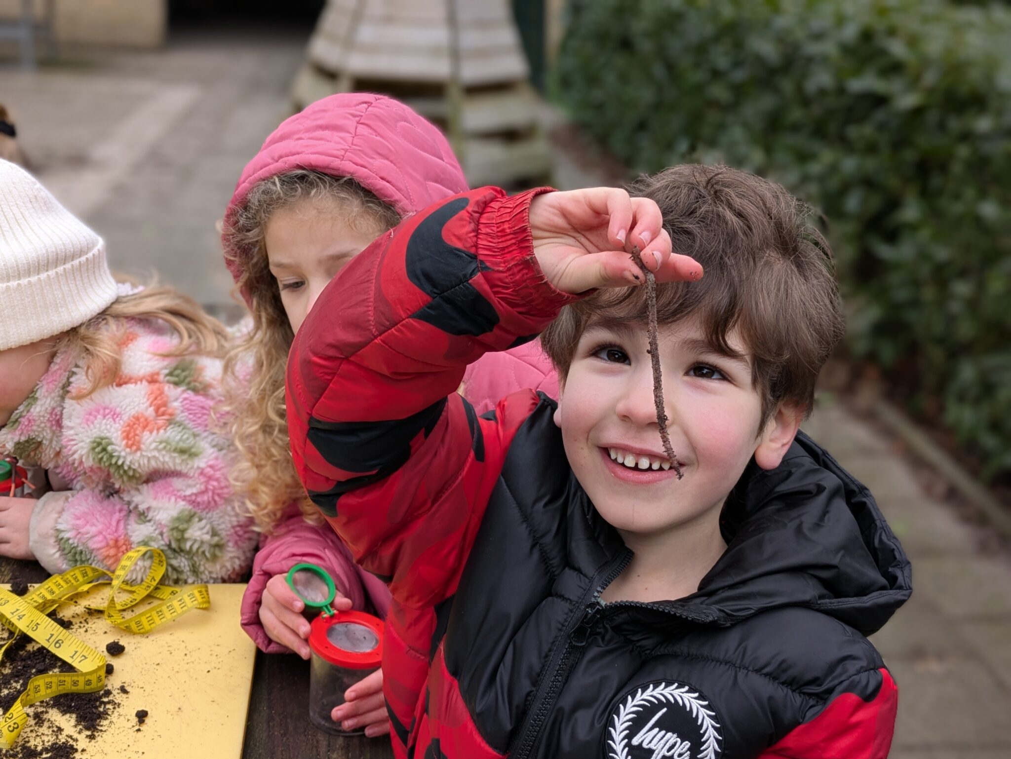 Een kind glimlacht terwijl hij een lange worm omhooghoudt; nog twee kinderen bekijken grond.
