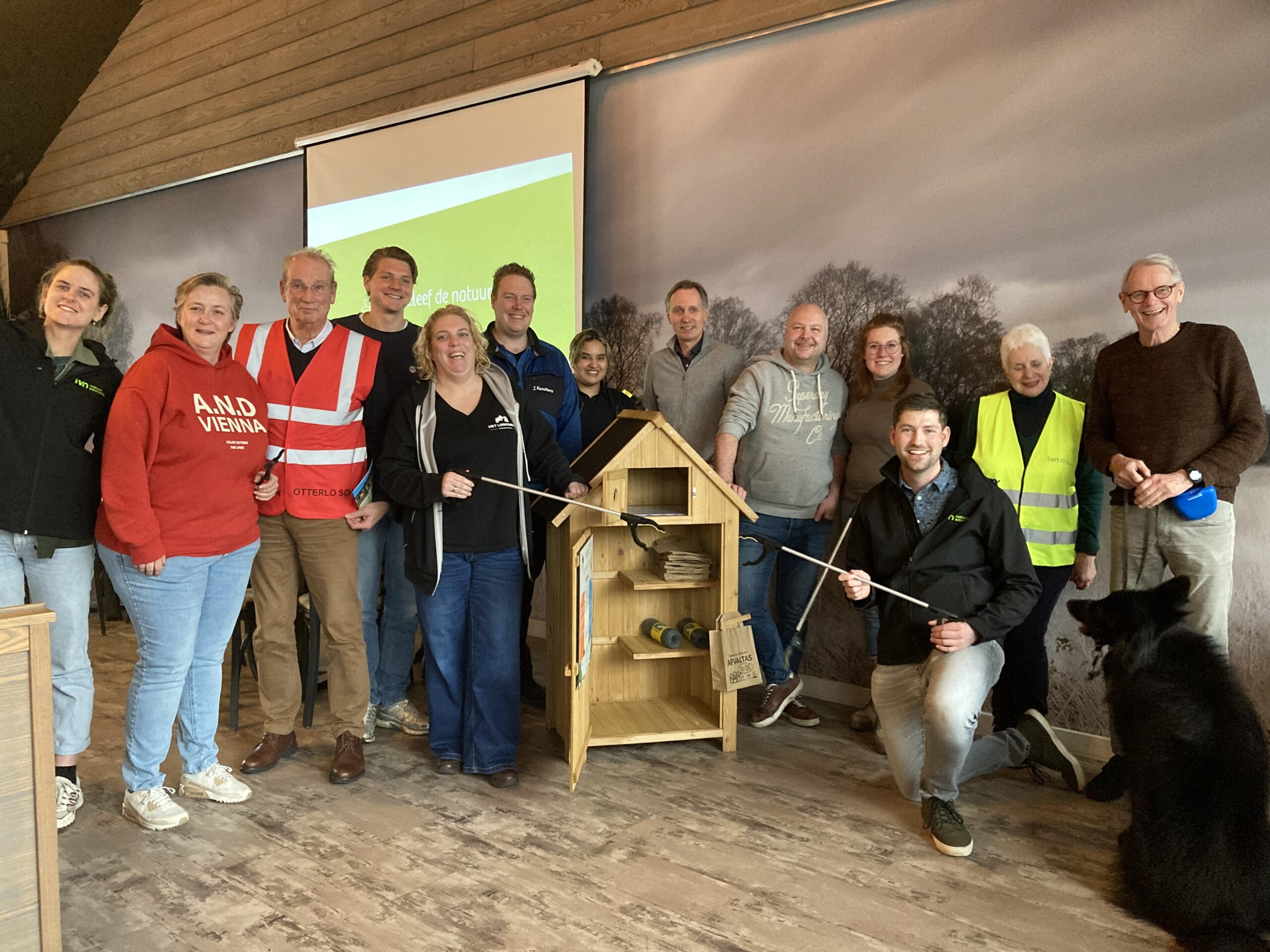 Groep mensen met zelfgemaakt houten insectenhotel, twee dragen veiligheidshesjes, grote zwarte hond erbij.