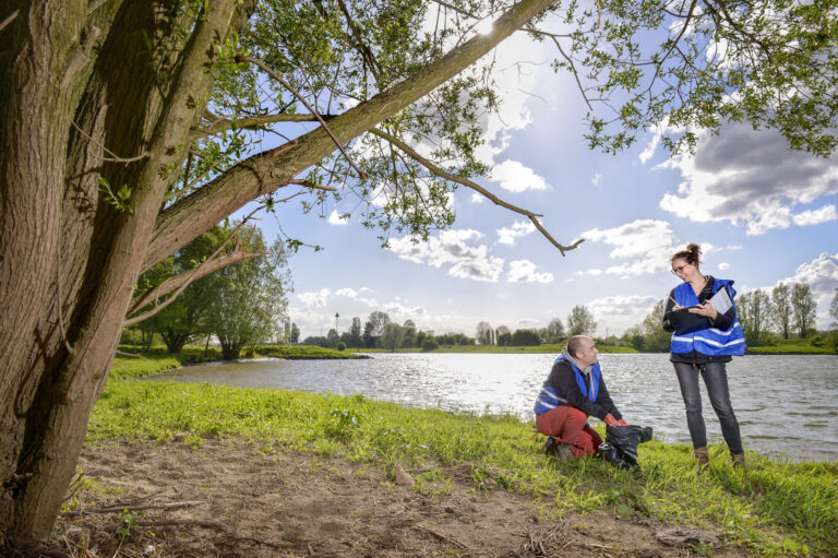 Twee mensen in blauwe veiligheidsvesten werken bij een rivier onder een boom op een zonnige dag.