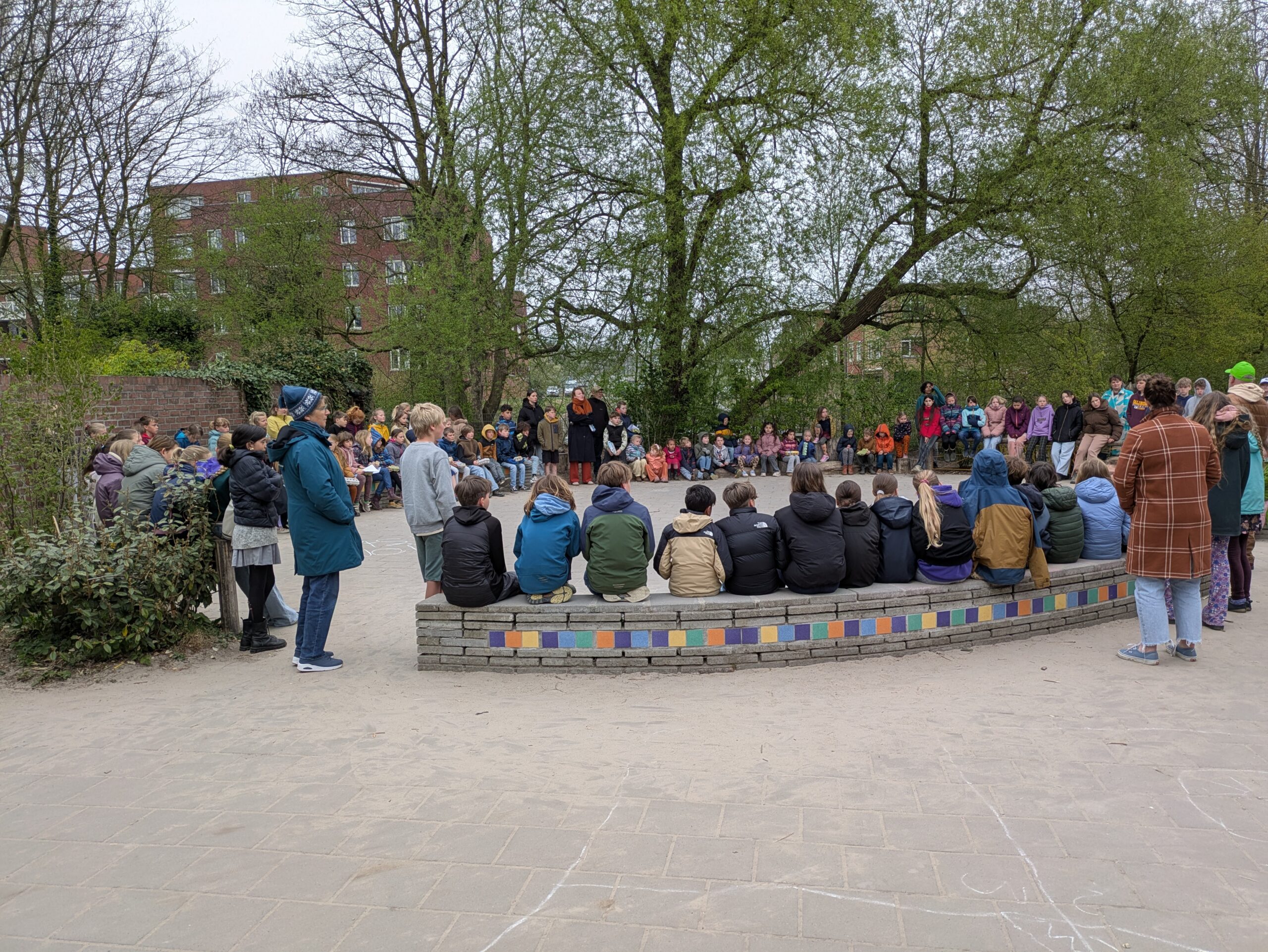 Groep mensen zit buiten in een cirkel op banken met kleurrijke mozaïektegels, omringd door bomen.