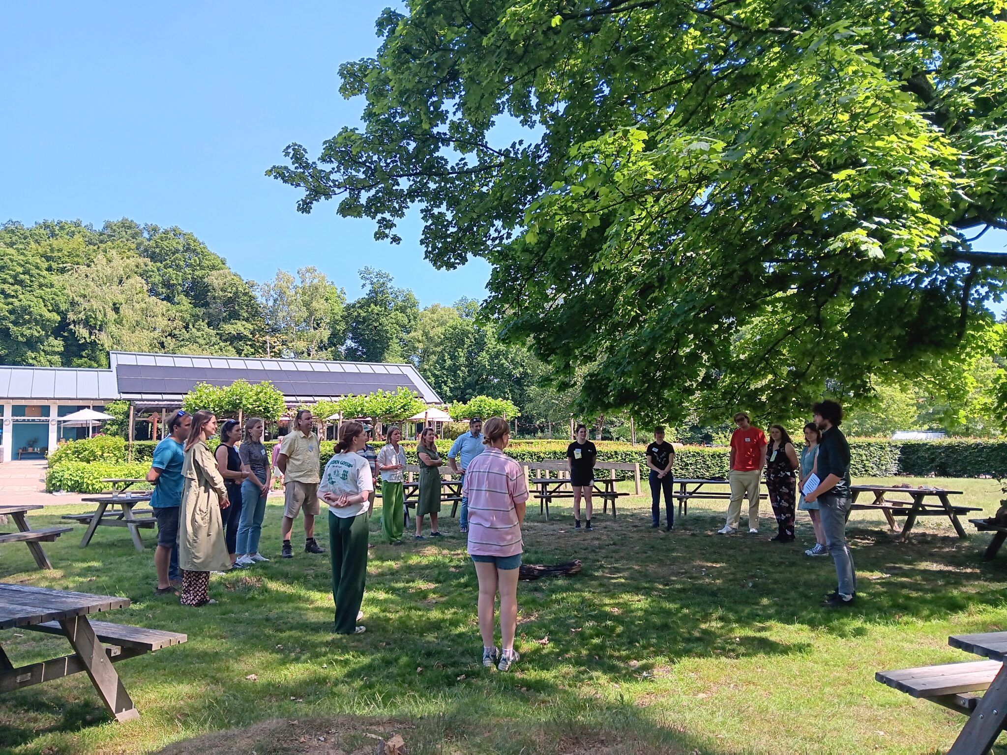 Mensen staan in een kring op een zonnige dag in een park met bomen en picknicktafels.