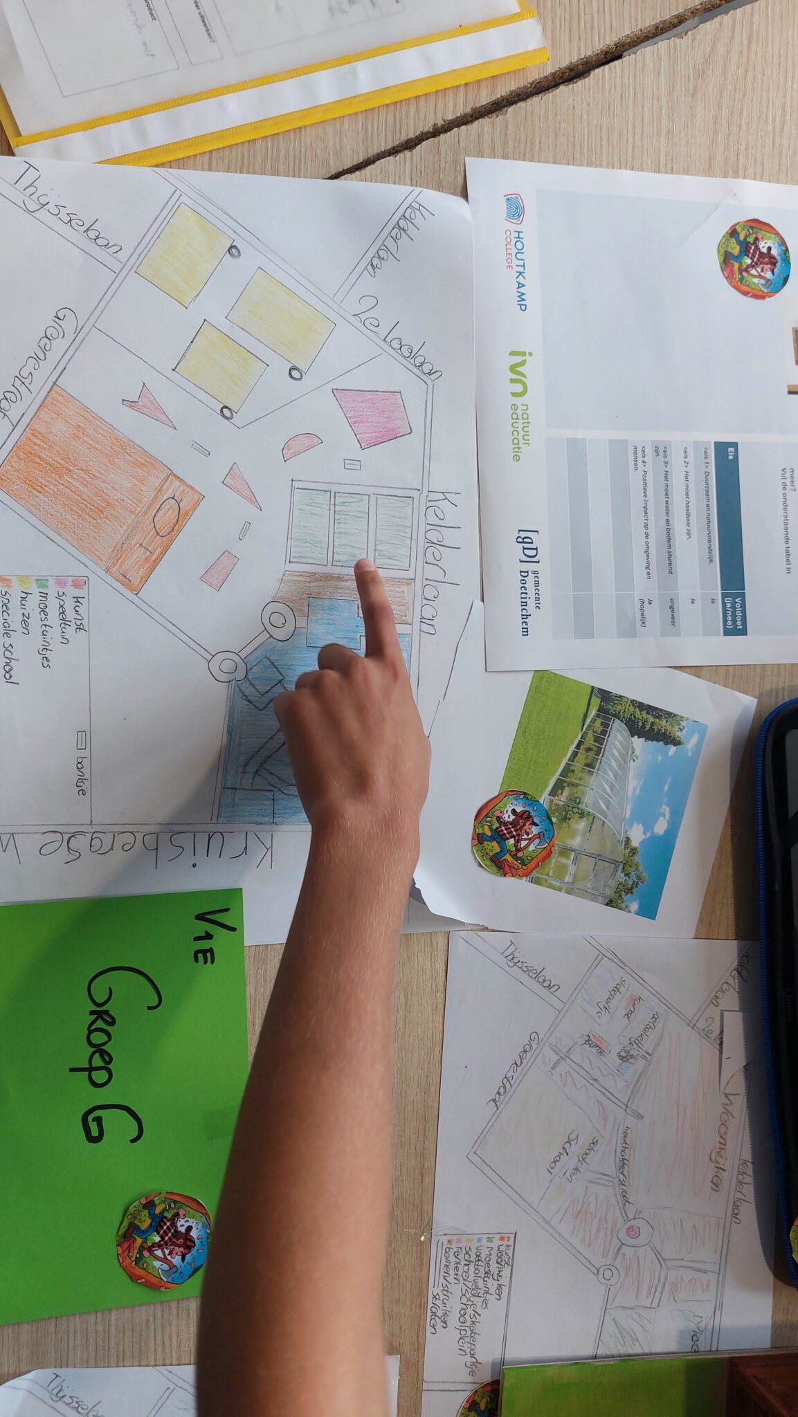 Een kind wijst naar een ingekleurde plattegrond op een tafel vol papieren en schoolwerk.