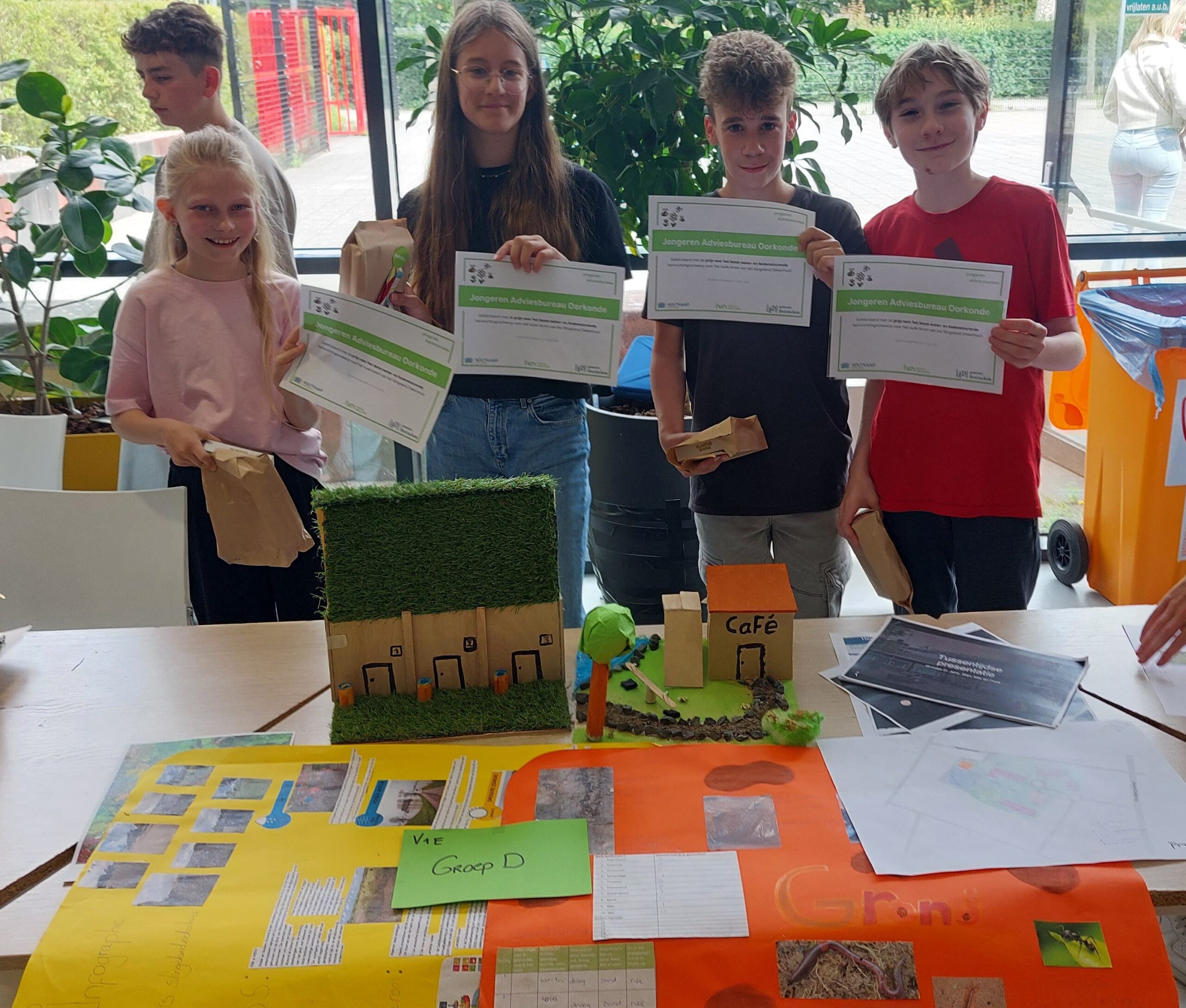 Vier kinderen tonen certificaten naast een maquette met grasdak en cafebord.
