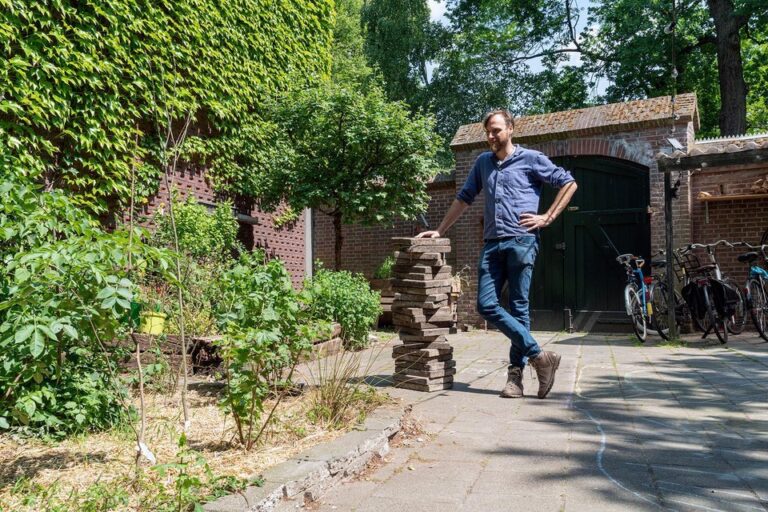 Man leunt op stapel stenen in zonnige tuin met klimop, fietsen en een schuurtje op de achtergrond.