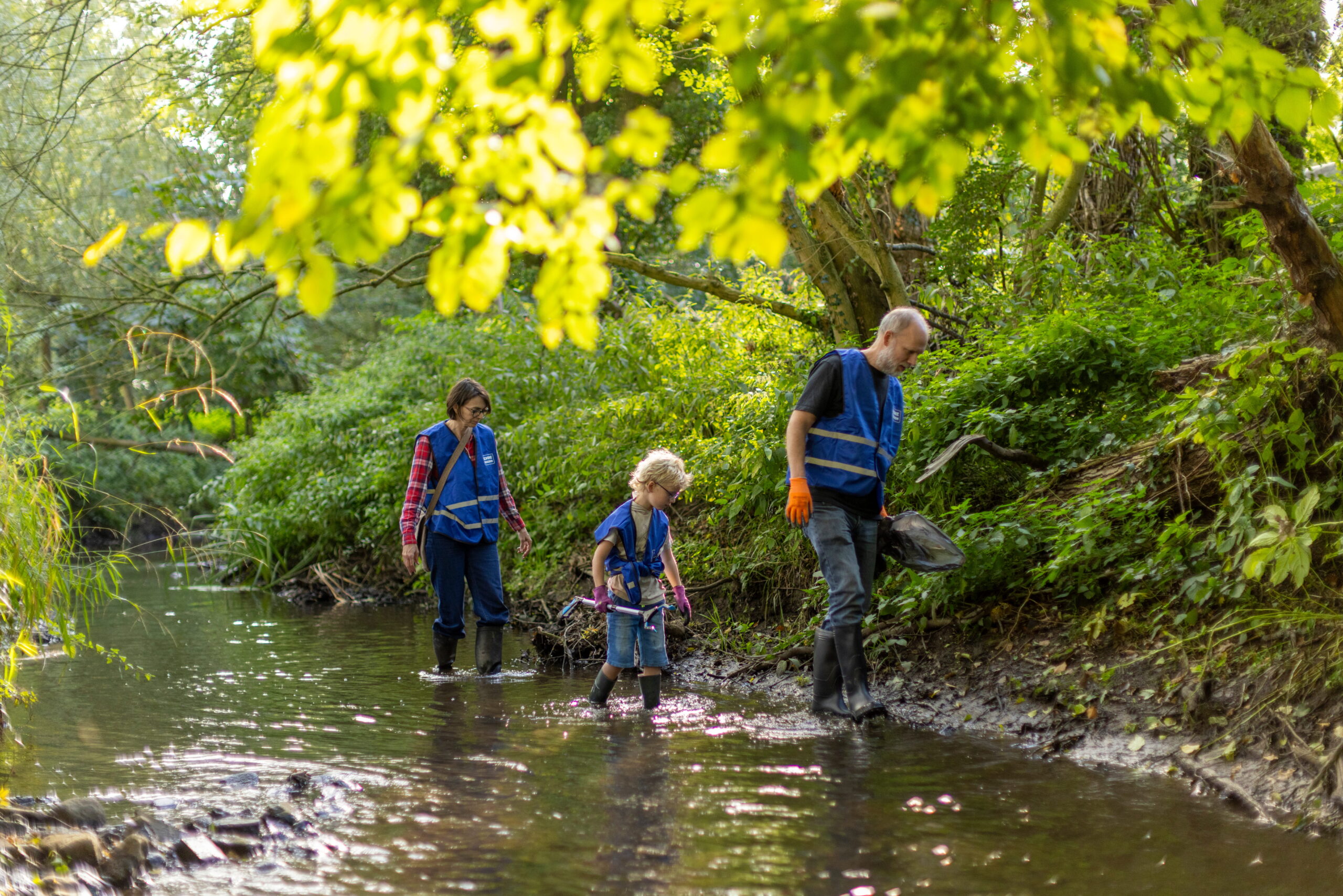 Drie mensen met blauwe hesjes lopen in een beek met groene bomen rondom.