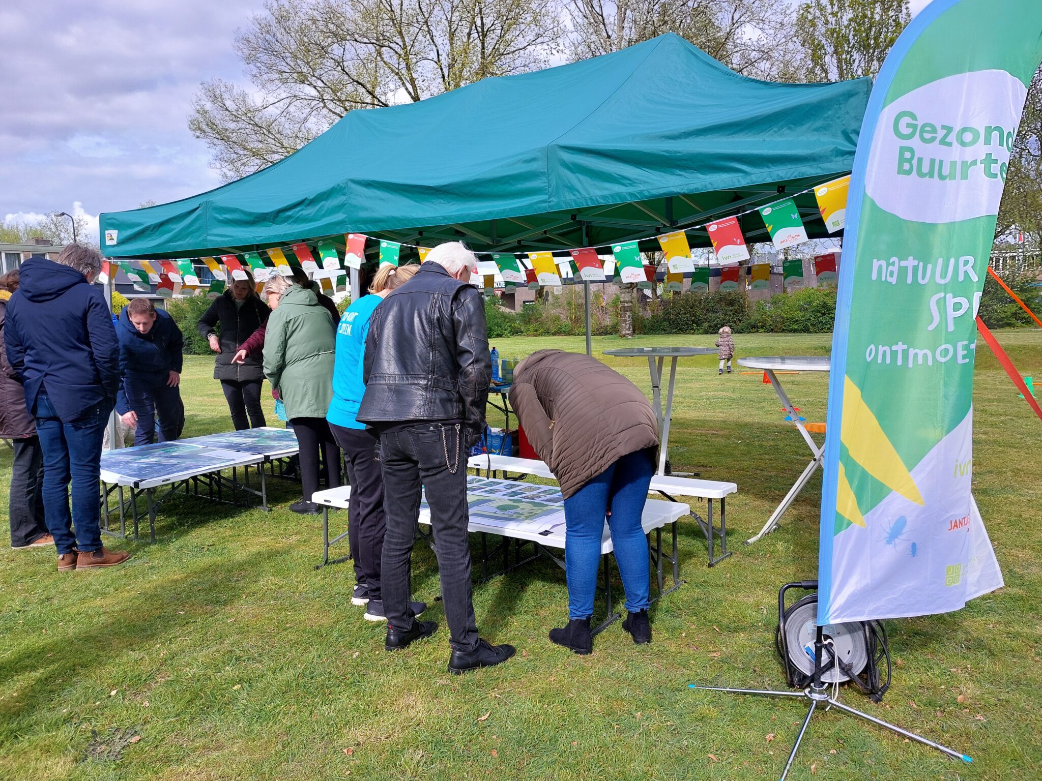 Mensen verzamelen zich onder een groene tent voor een buurtactiviteit in een park.