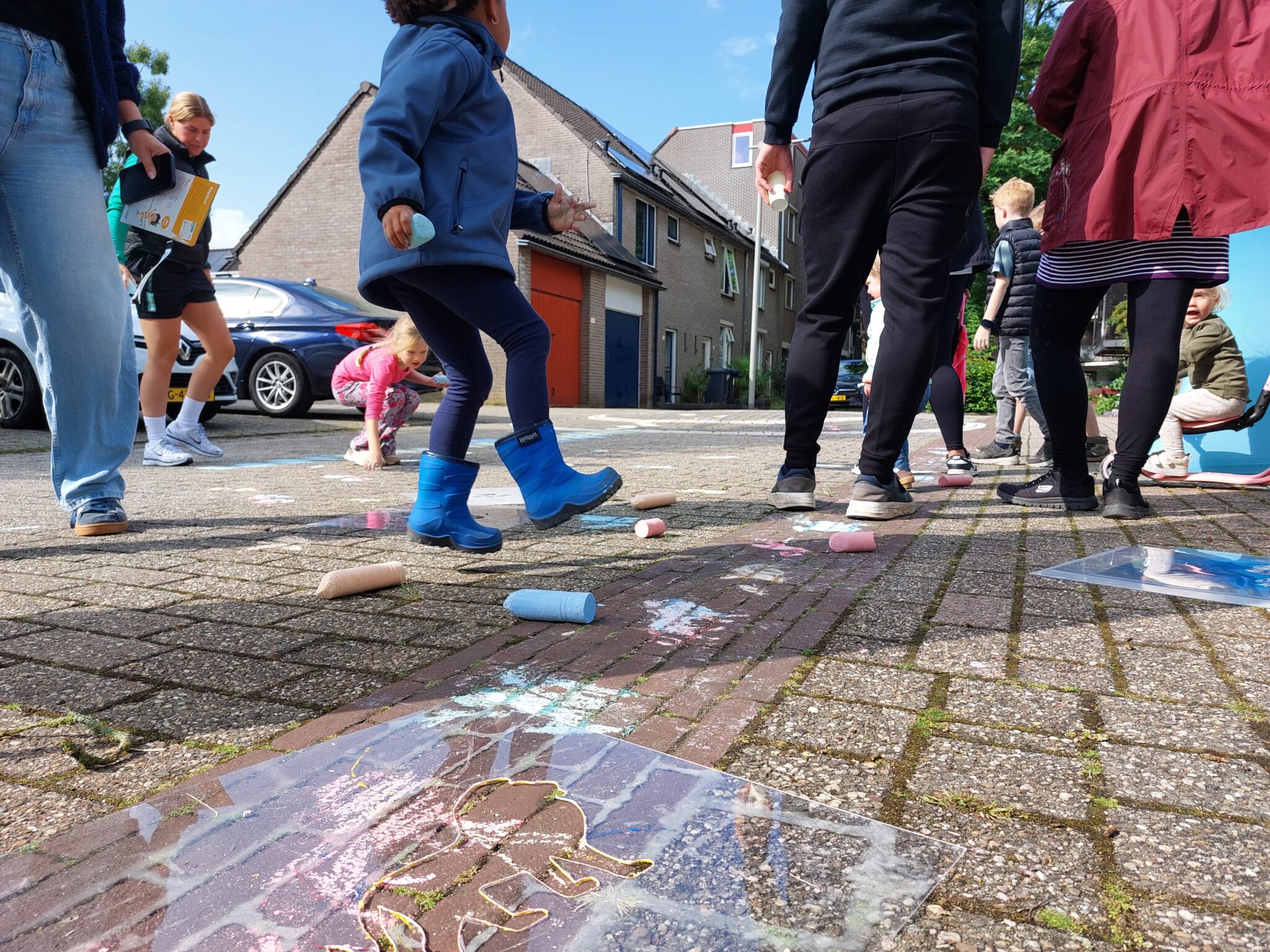 Kinderen tekenen met stoepkrijt op straat; volwassenen kijken toe in een woonwijk.