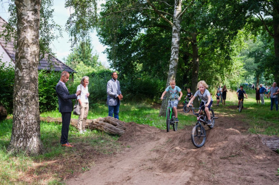 Kinderen fietsen op een bospad terwijl volwassenen toekijken in een groene omgeving.