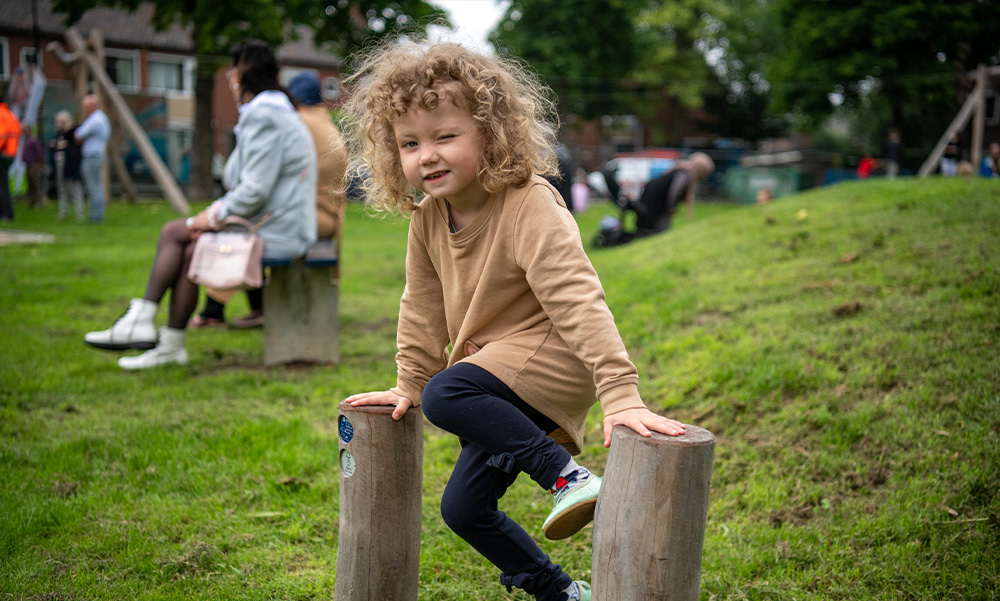 Kind speelt buiten op houten paal in park; mensen op achtergrond.