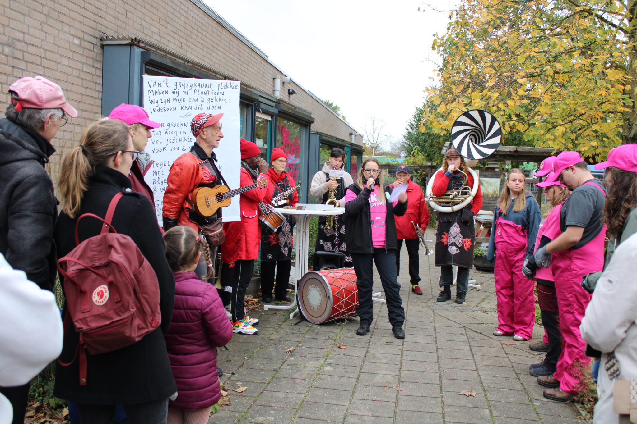 Muzikanten en deelnemers in roze kleding tijdens een buitenevenement met een spandoek en muzikale optredens.
