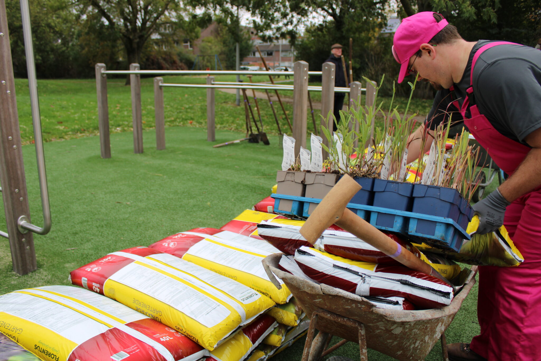 Man met roze werkkleding legt plantenbakken in een kruiwagen naast stapels zakken potgrond.