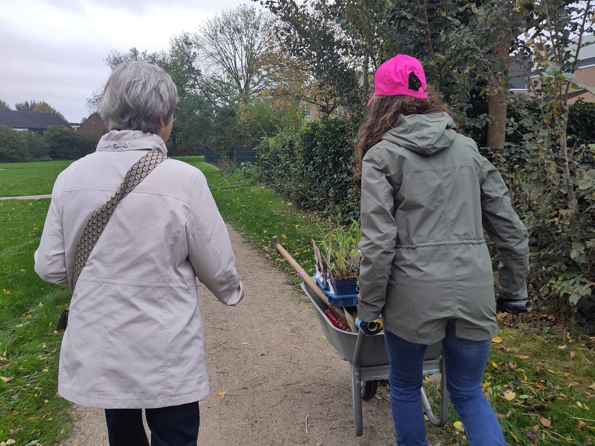 Twee personen wandelen op een bospad; één duwt een kruiwagen met planten.