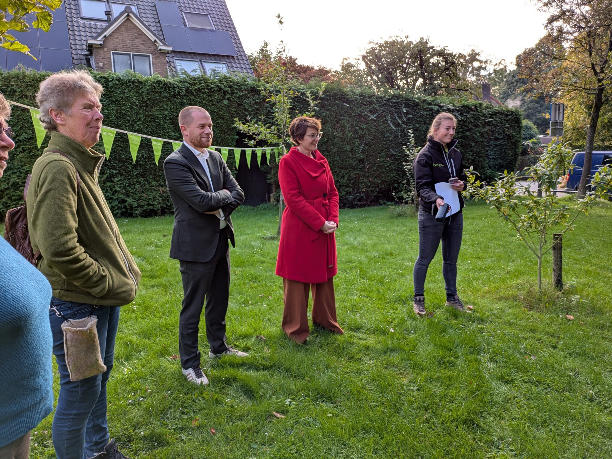 Mensen staan buiten in een tuin bij een jonge boom, met feestelijke vlaggetjes op de achtergrond.