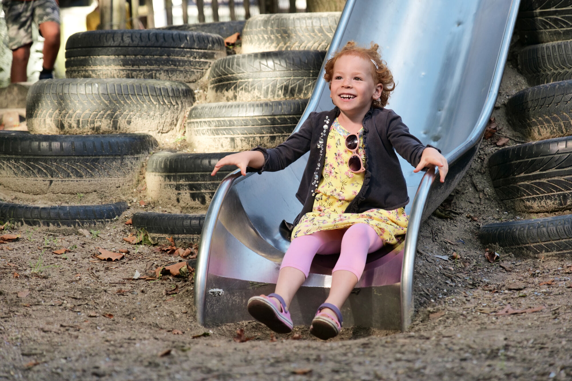 Kind glijdt blij van een metalen glijbaan omgeven door gestapelde autobanden in speeltuin.
