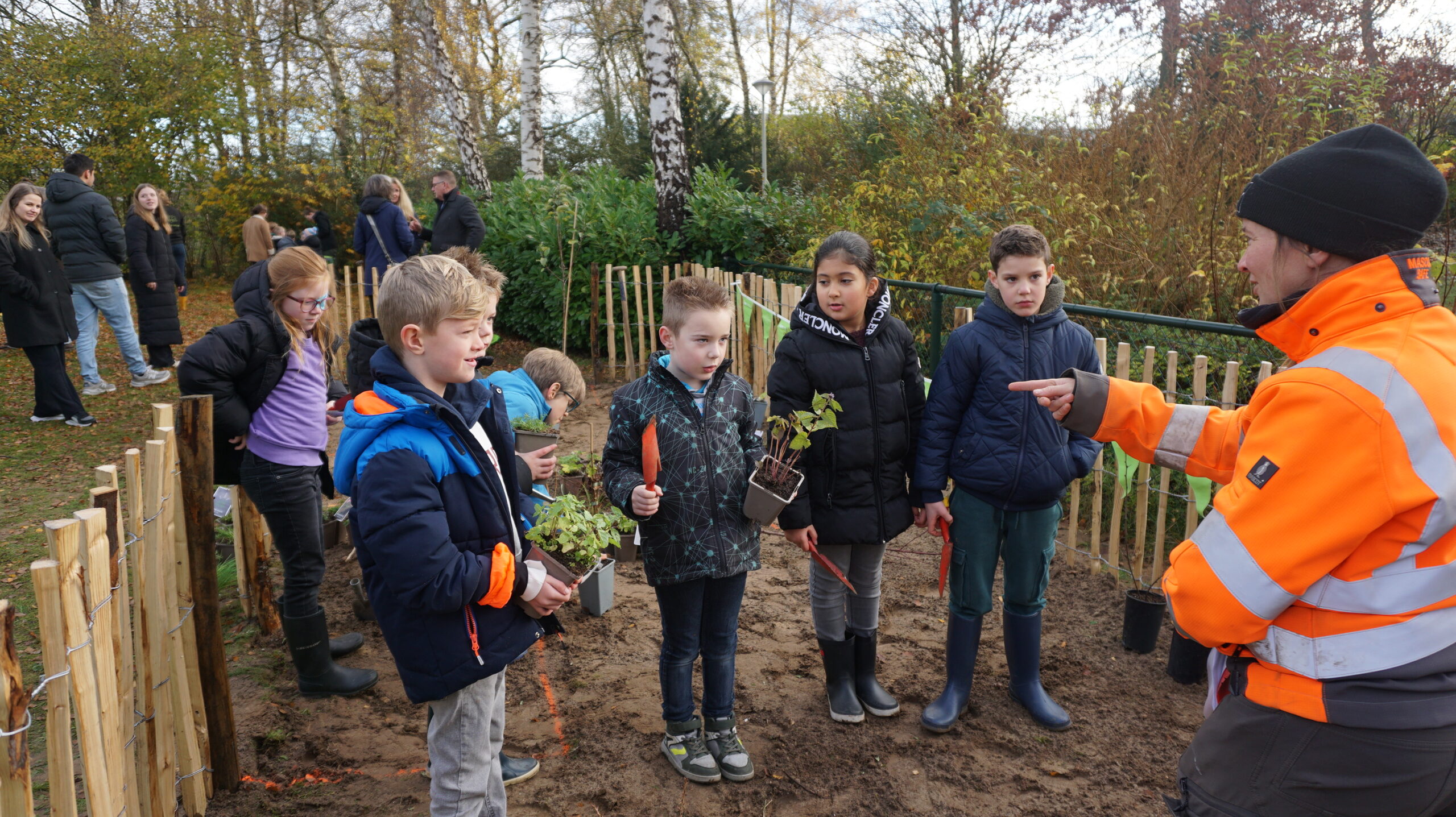 Kinderen planten jonge boompjes met begeleiding van een volwassene in een oranje jas.