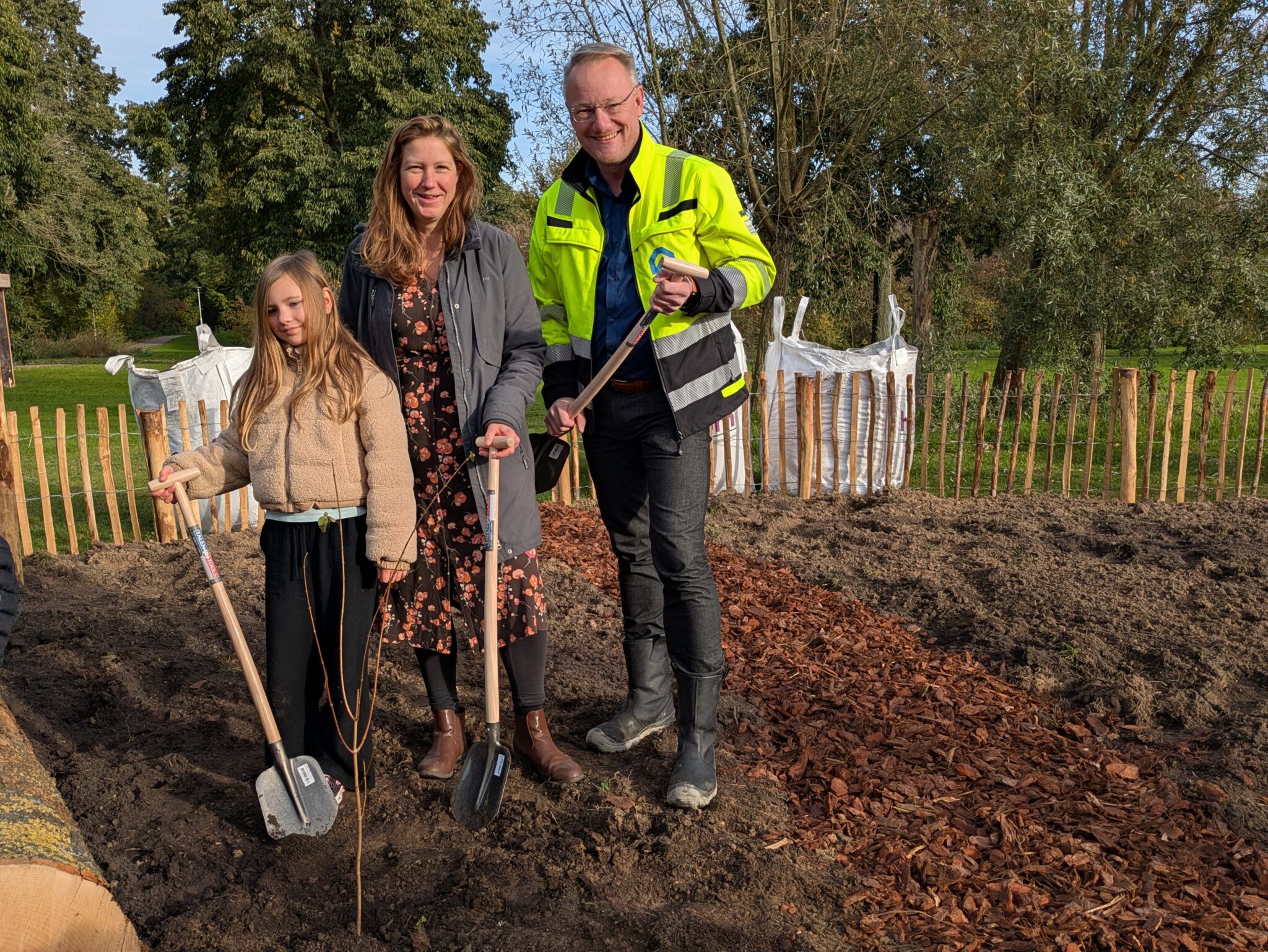 Een man, vrouw en kind planten een boom in een tuin met schoppen.