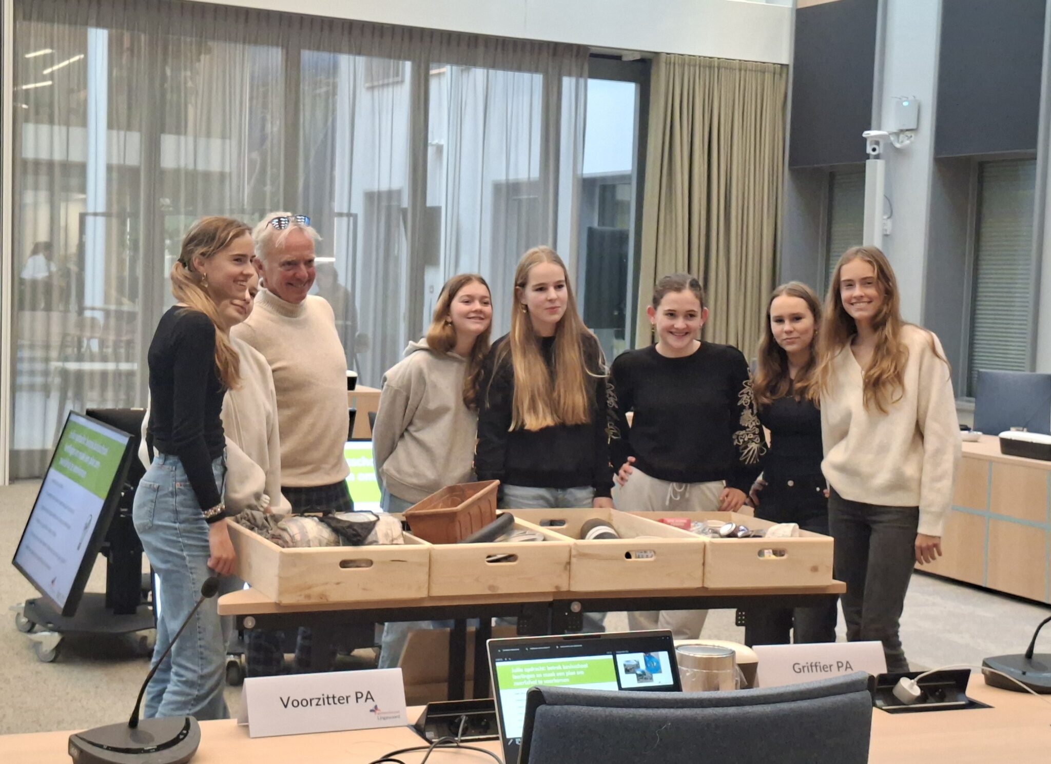 Groep mensen poseert voor houten kistjes in vergaderruimte met naamborden en schermen.