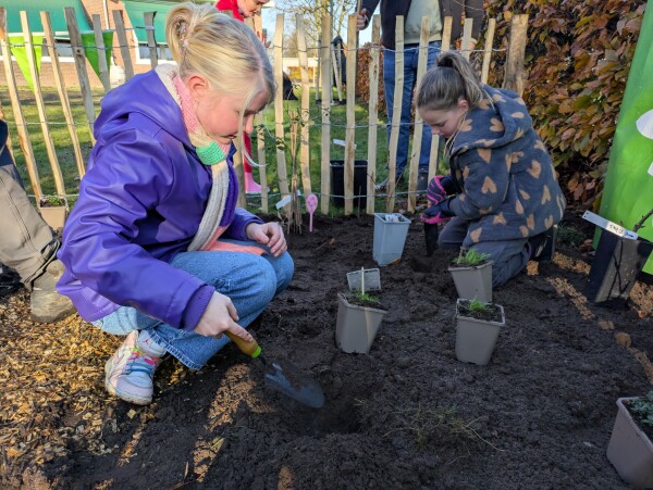 Kinderen planten bloemen in een tuin met potjes en schepjes, naast een houten hek.