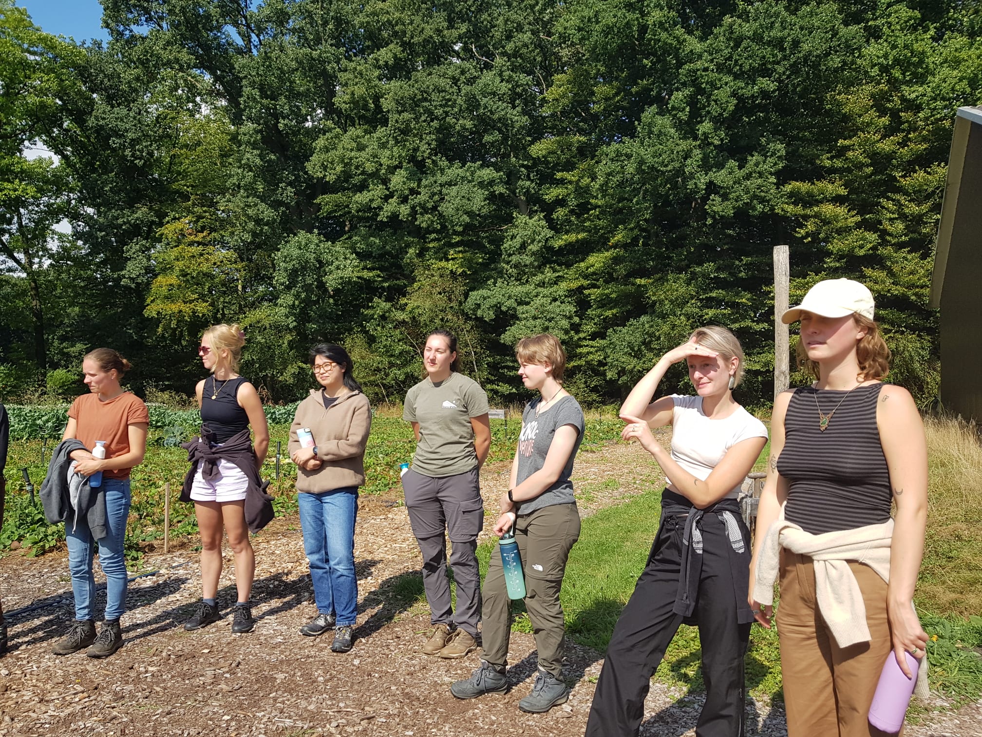 Groep mensen staat buiten in zonlicht met bomen op de achtergrond. Ze kijken dezelfde kant op.
