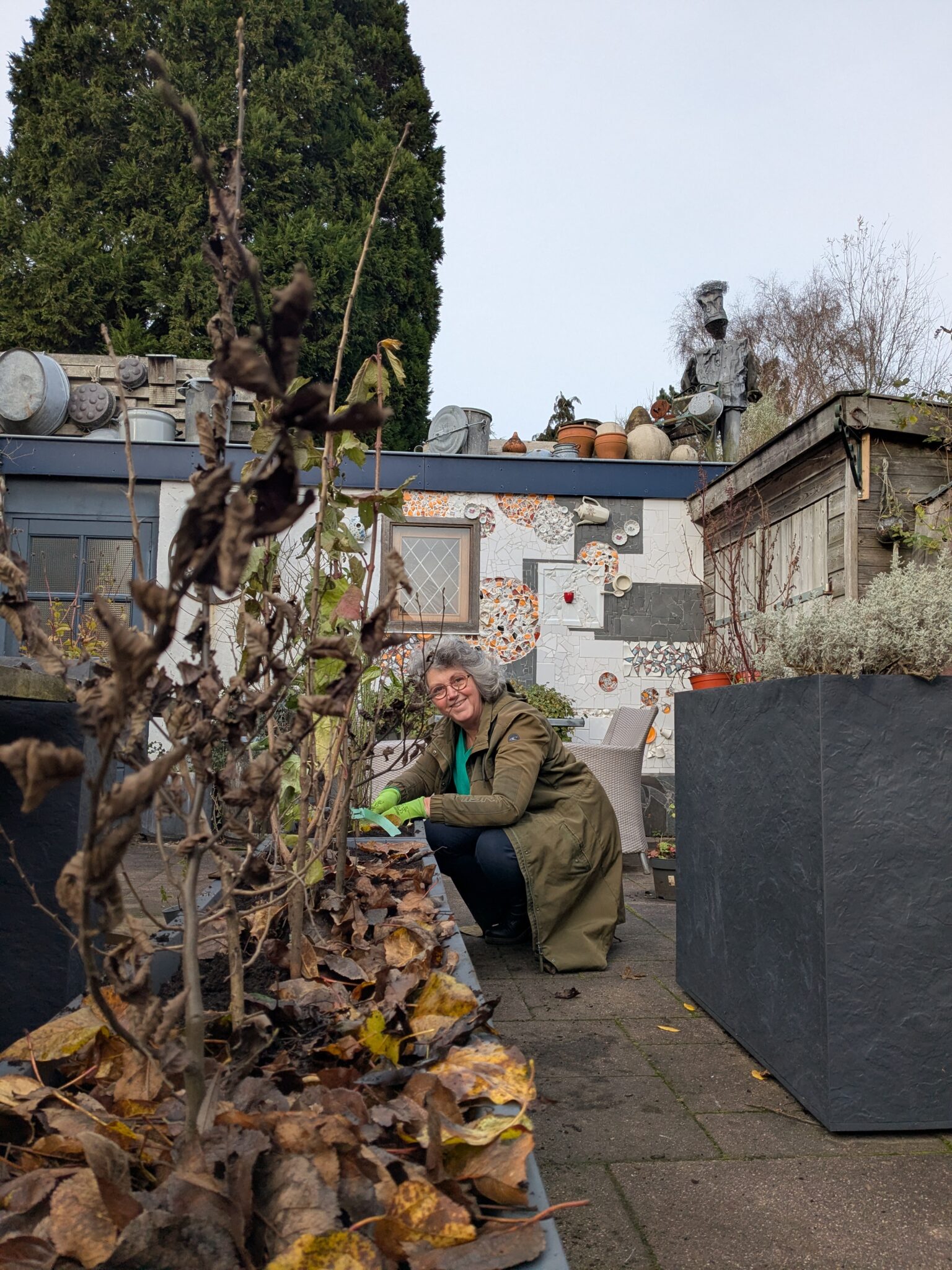 Vrouw in een tuin bij een verhoogde plantenbak met verdorde bladeren, omgeven door decoratieve objecten.