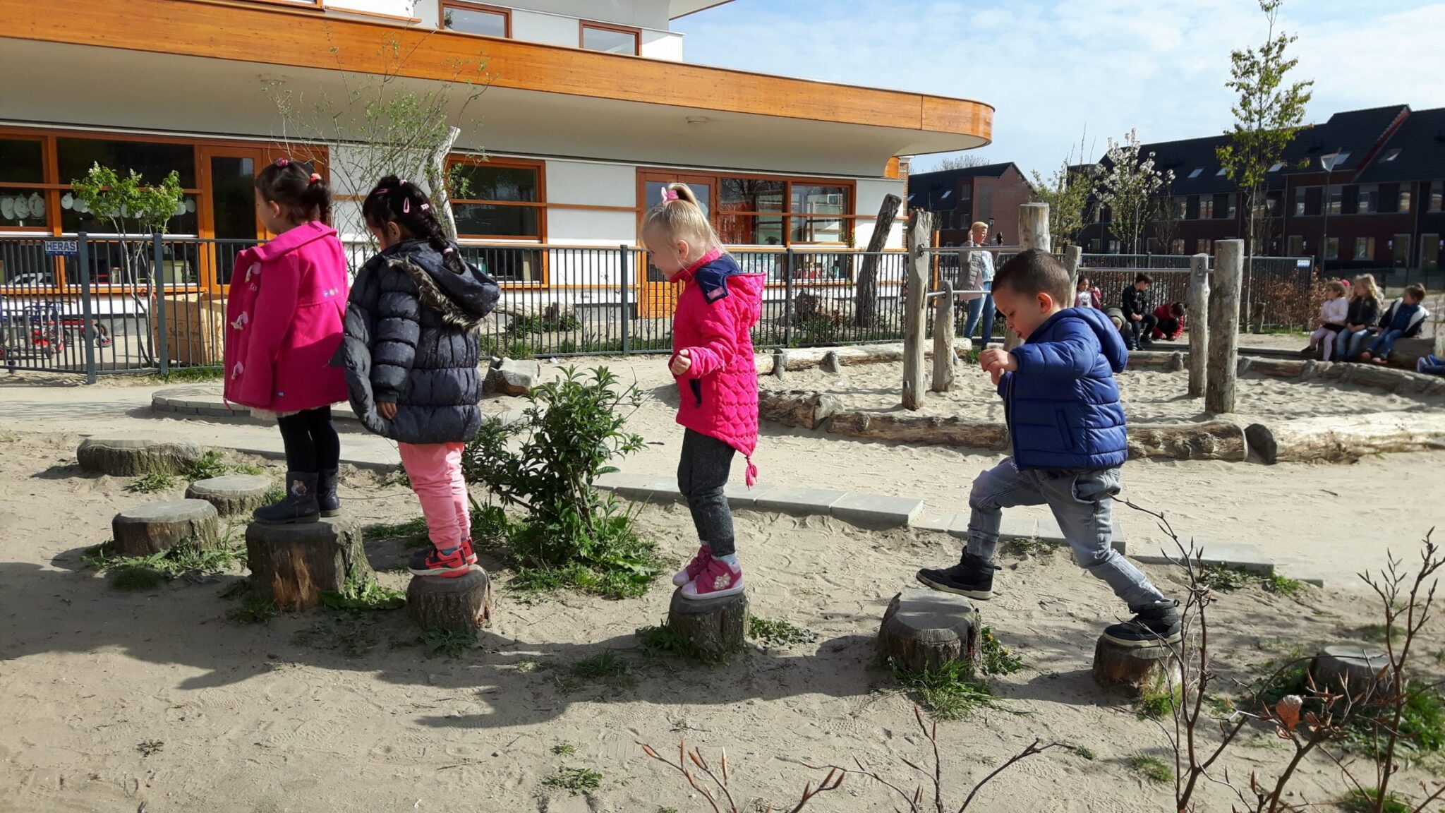 Kinderen lopen over boomstammen op een zandige speelplaats bij een gebouw met houten details.
