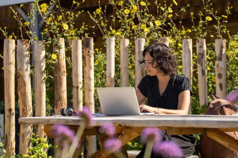 Vrouw werkt buiten op laptop aan houten tafel, omringd door bloemen en houten hek.