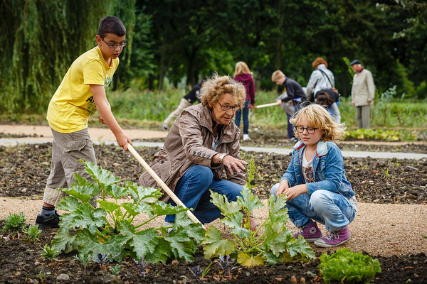 Drie mensen werken samen in een tuin; een oudere vrouw en twee kinderen inspecteren planten.