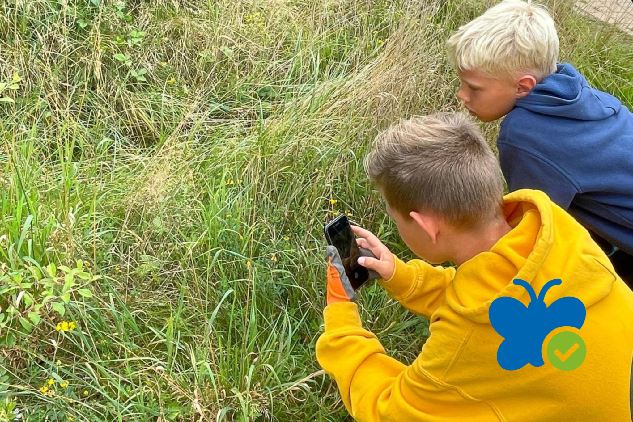Kinderen onderzoeken gras met telefoon; een draagt een gele hoodie met een vlinderlogo.
