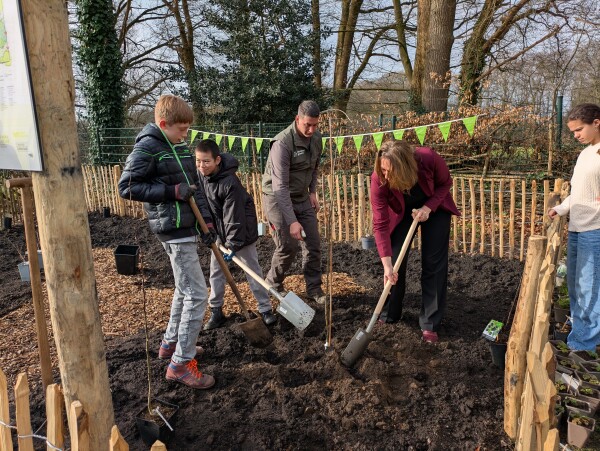Groep mensen graaft met schoppen in een tuin omringd door een houten hek en feestelijke vlaggetjes.
