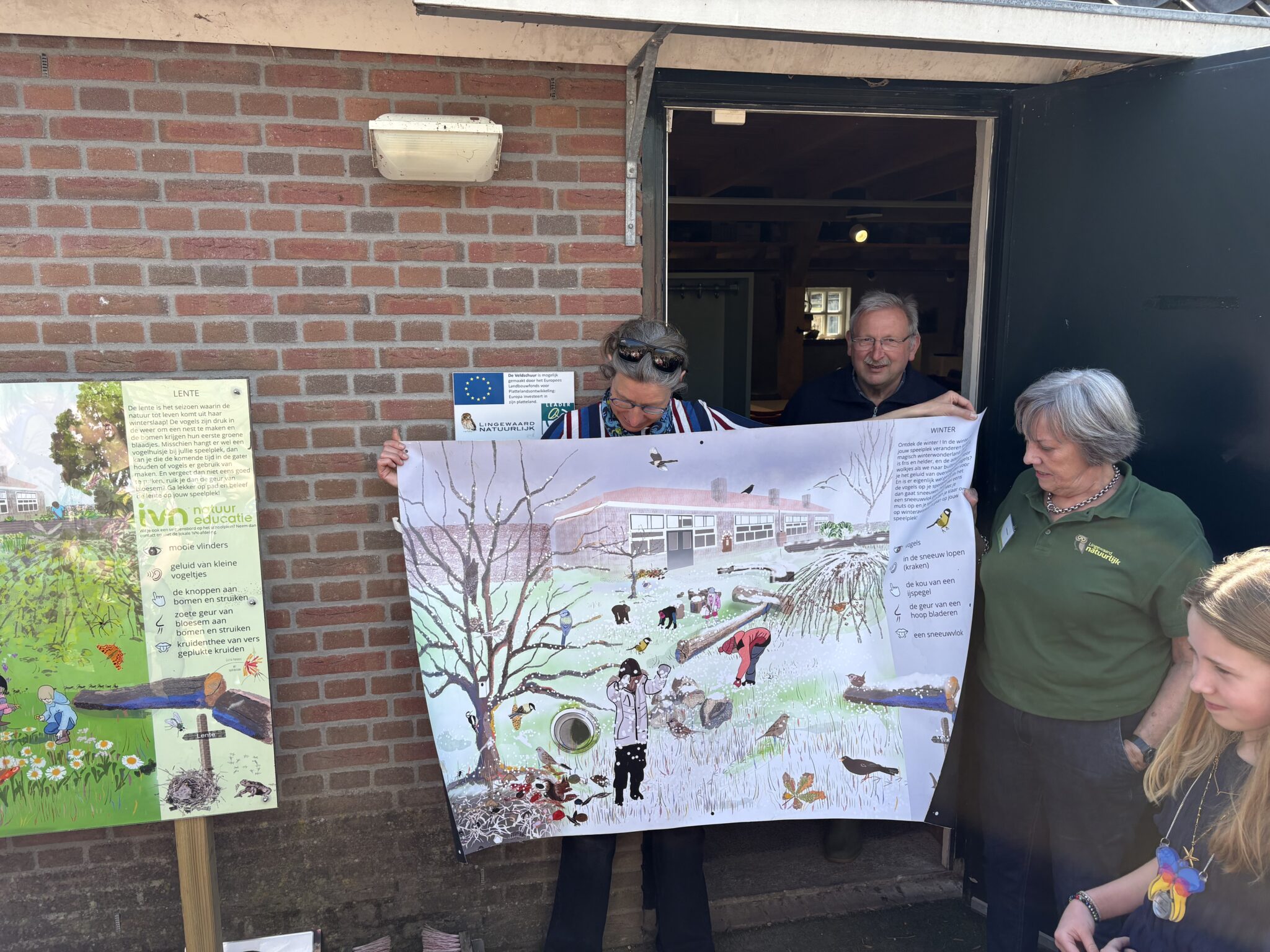 Mensen tonen een grote natuureducatieposter voor een gebouw, naast een informatiebord.