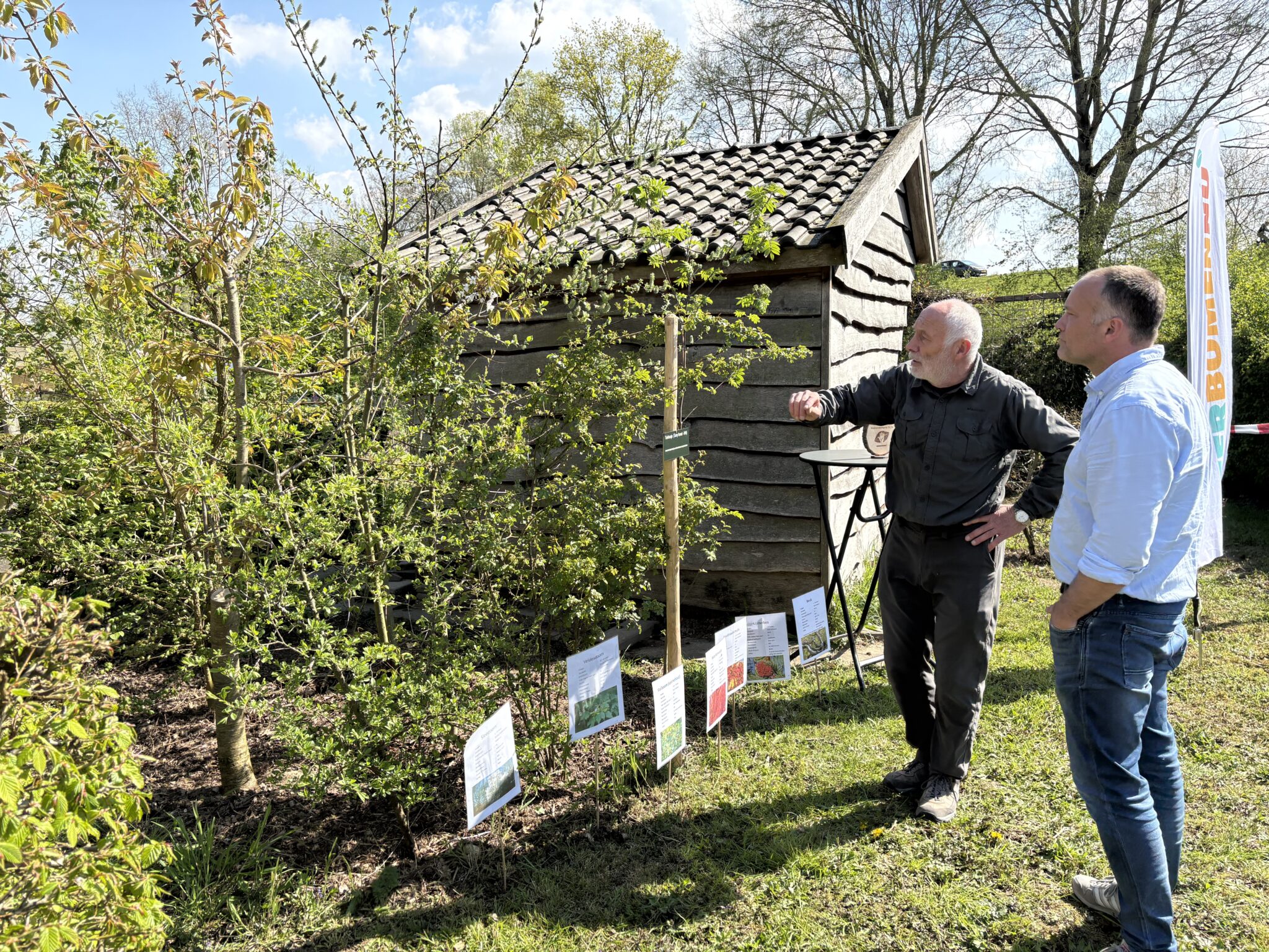 Twee mannen bekijken jonge bomen naast een houten schuurtje met informatieborden in een tuin.