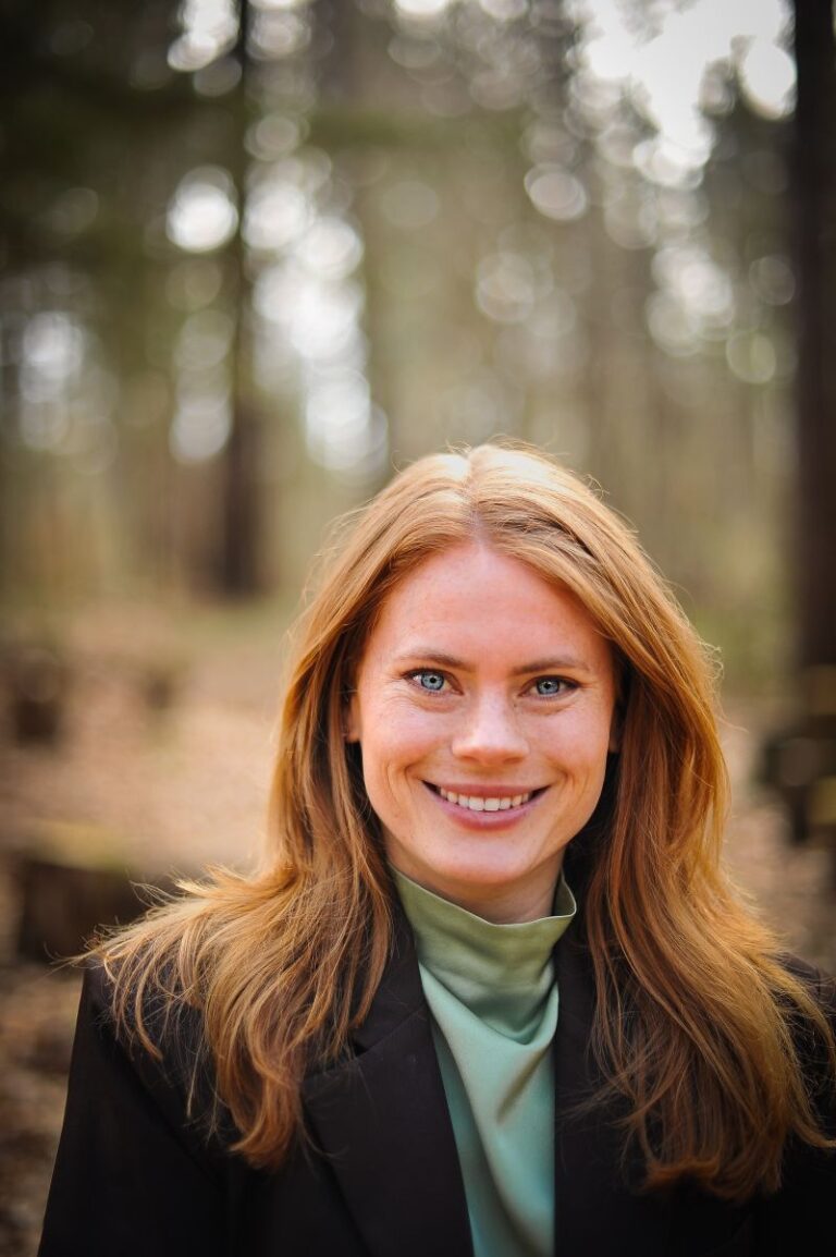 Vrouw glimlacht in een herfstbos, met onscherpe bomen op de achtergrond.