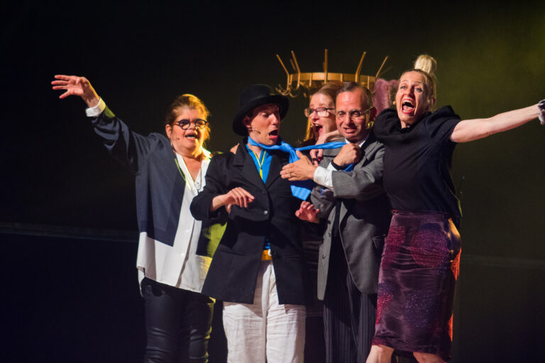 Groep acteurs op podium, lachen en dramatisch poseren met kleurrijke kleding en accessoires.