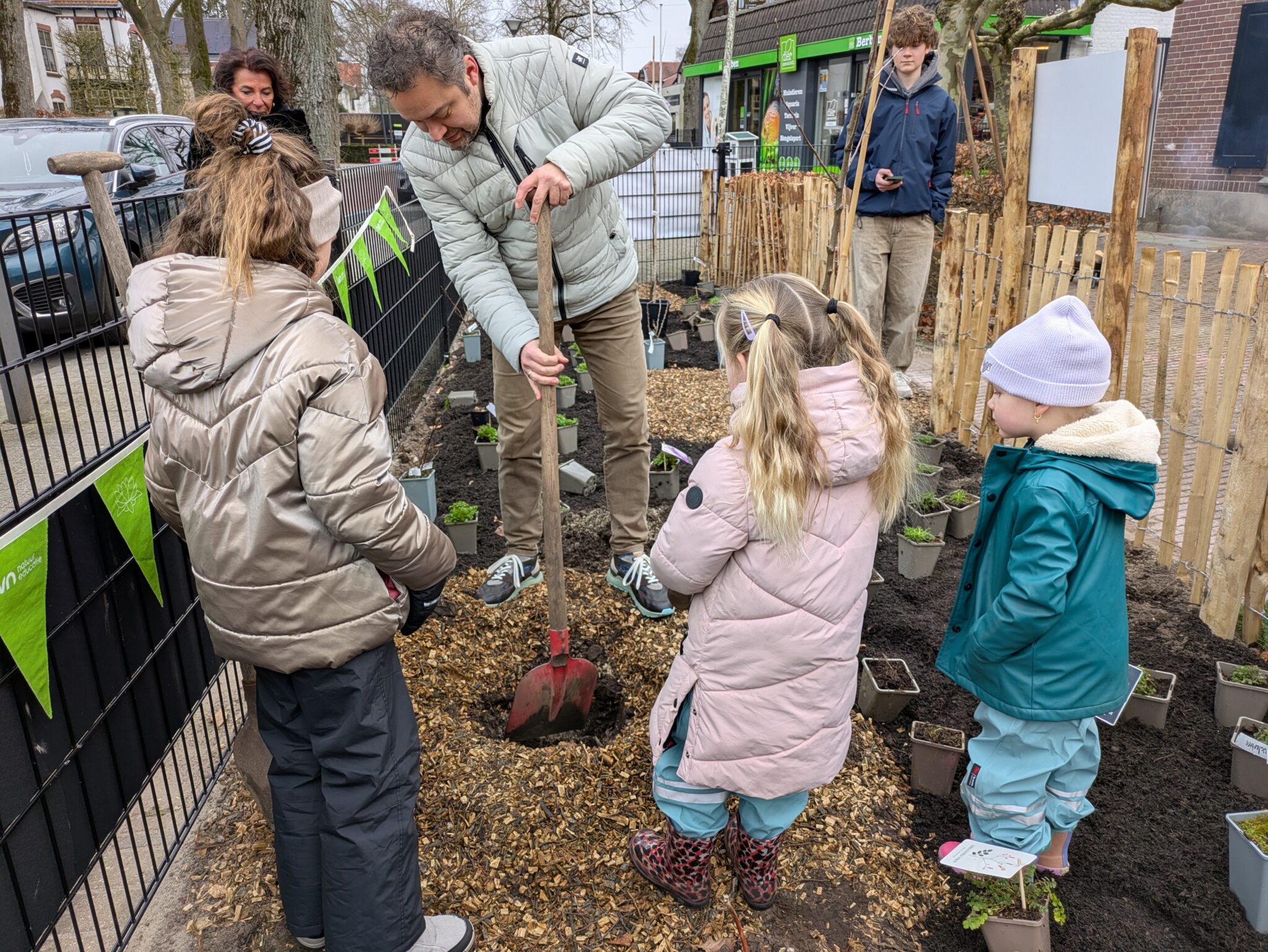 Volwassenen en kinderen planten samen zaailingen in een tuin met hek en houten omheining.