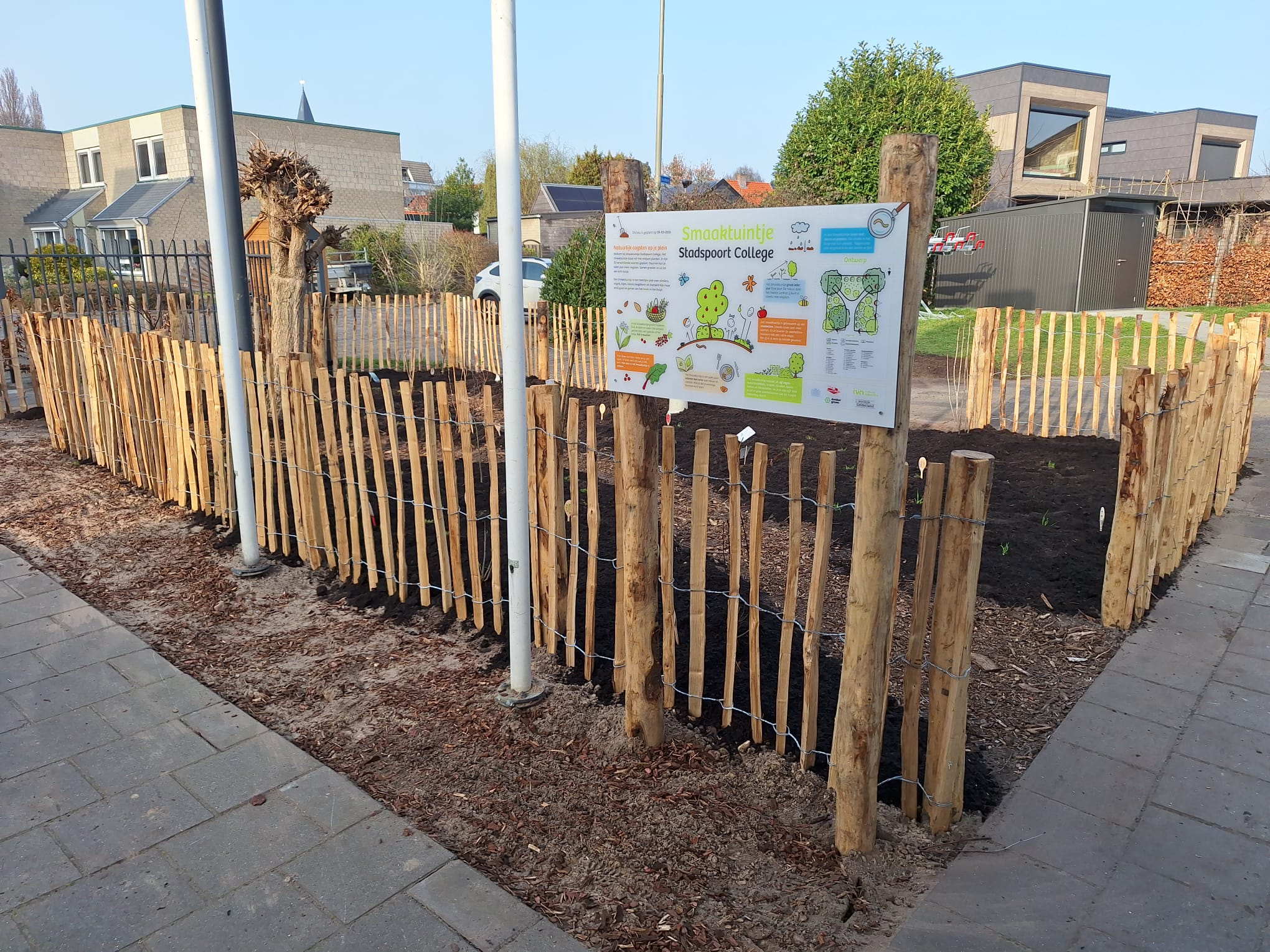 Kleurrijk infobord bij een tuin binnen een houten hekwerk naast een schoolgebouw.