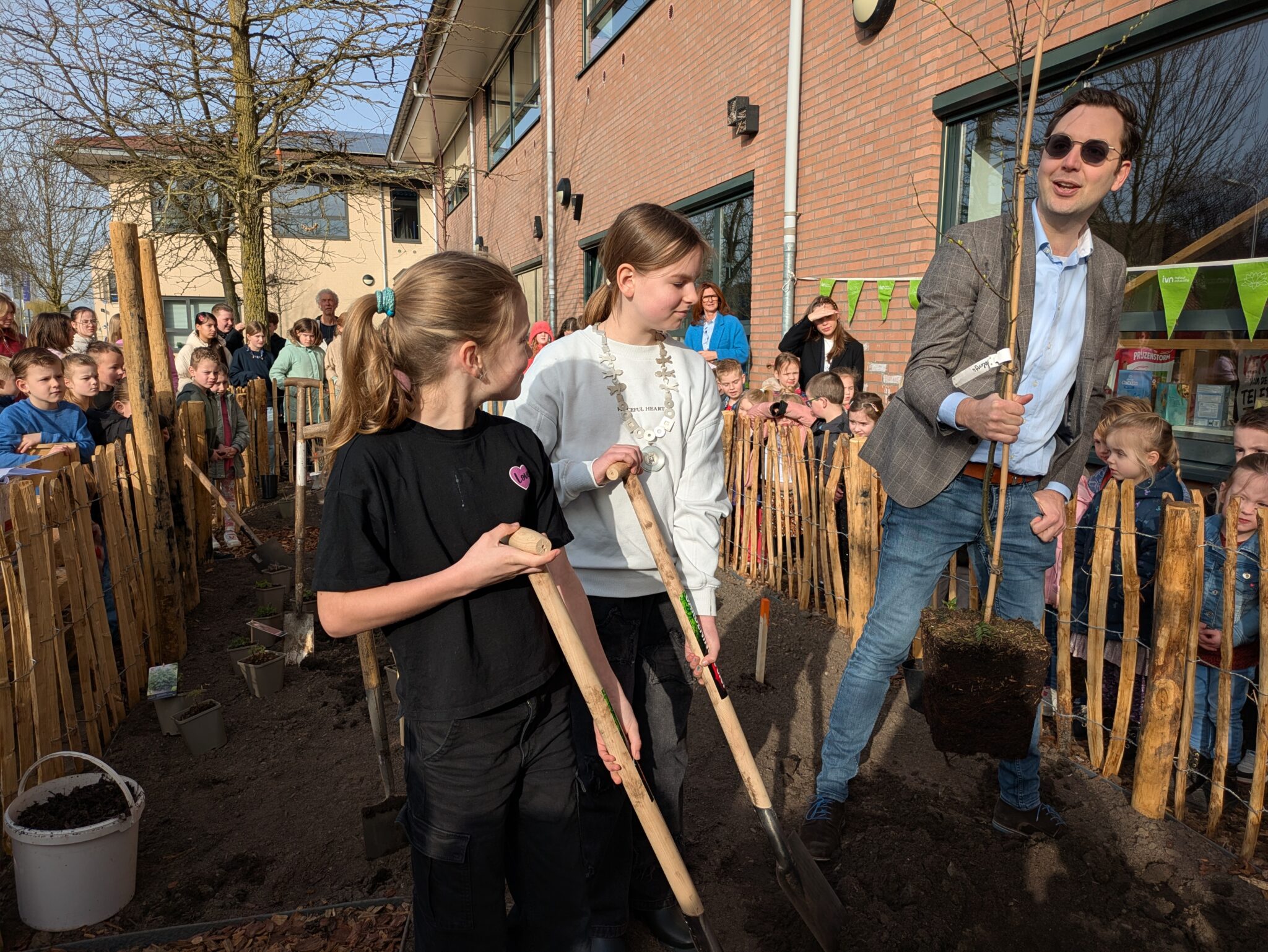 Mensen planten een boom met kinderen op een schoolplein, omringd door toeschouwers en hekwerk.