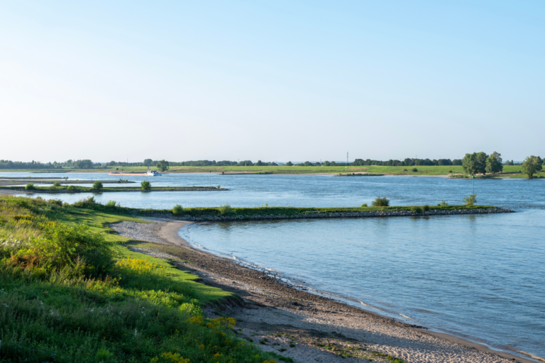 Rivier met graslanden en zandstrand bij zonsondergang, met groene oevers en een boot in de verte.