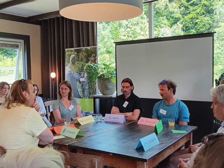 Groepsdiscussie in vergaderruimte, met zes deelnemers rond tafel en een leeg projectiescherm.