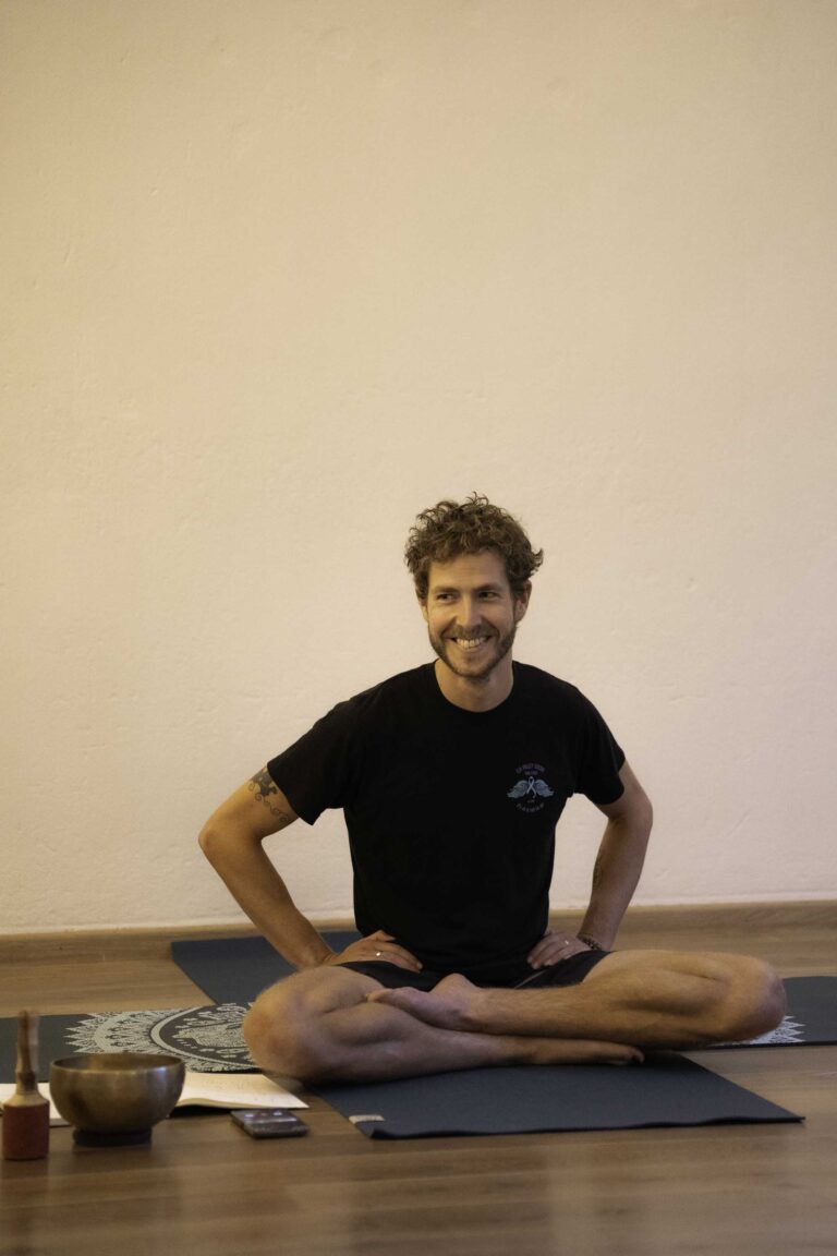 Man zit op een yogamat, glimlachend, met een klankschaal en boek naast hem.