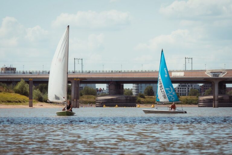 Twee zeilboten op een rivier, met een brug en gebouwen op de achtergrond.