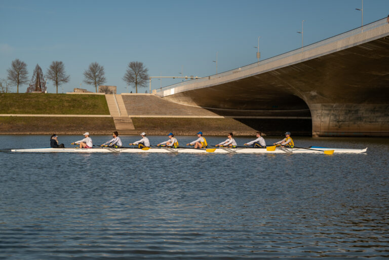 Roeiteam op rivier nabij brug op een zonnige dag, met kale bomen op de achtergrond.