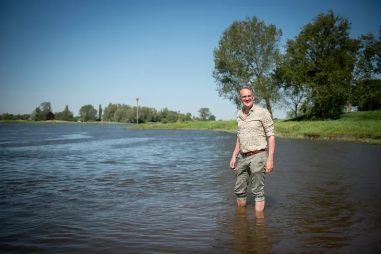 Een man staat glimlachend in een rivier met bomen op de achtergrond onder een blauwe hemel.