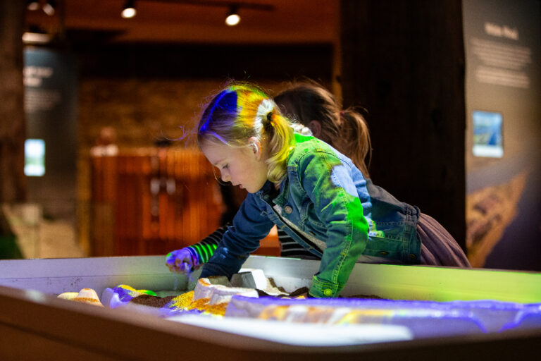 Een meisje speelt met zand in een interactieve zandbak, verlicht door kleurrijke projecties.