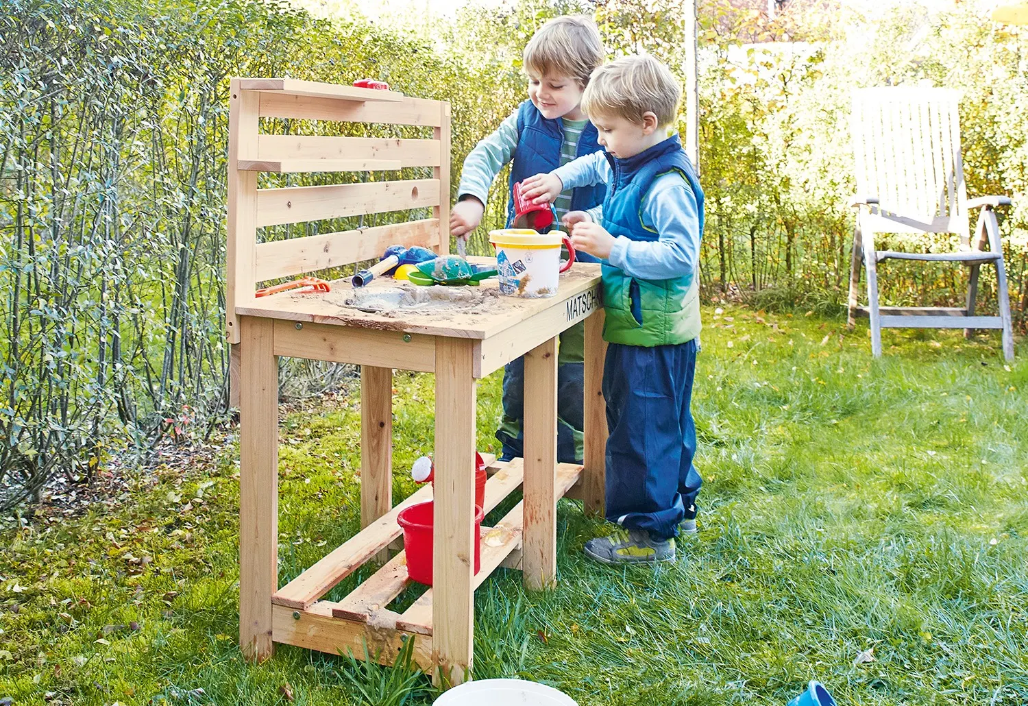 Twee kinderen spelen met zand en speelgoed aan een houten speelkeukentje in een grasrijke tuin.