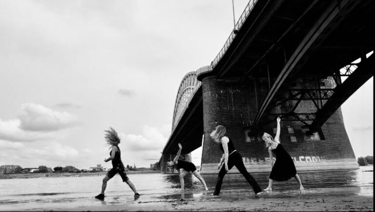 Vier mensen dansen onder een grote brug aan het water. Het is een zwart-wit afbeelding.