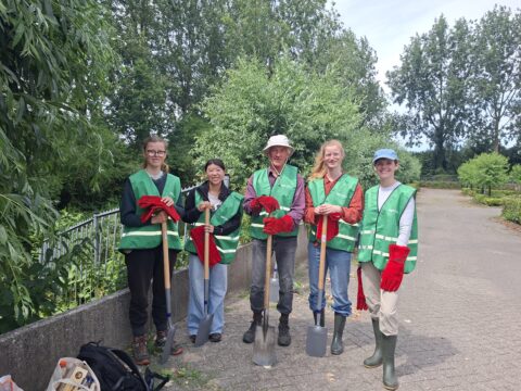 Vijf mensen in groene hesjes met scheppen en handschoenen, omringd door bomen in een park.
