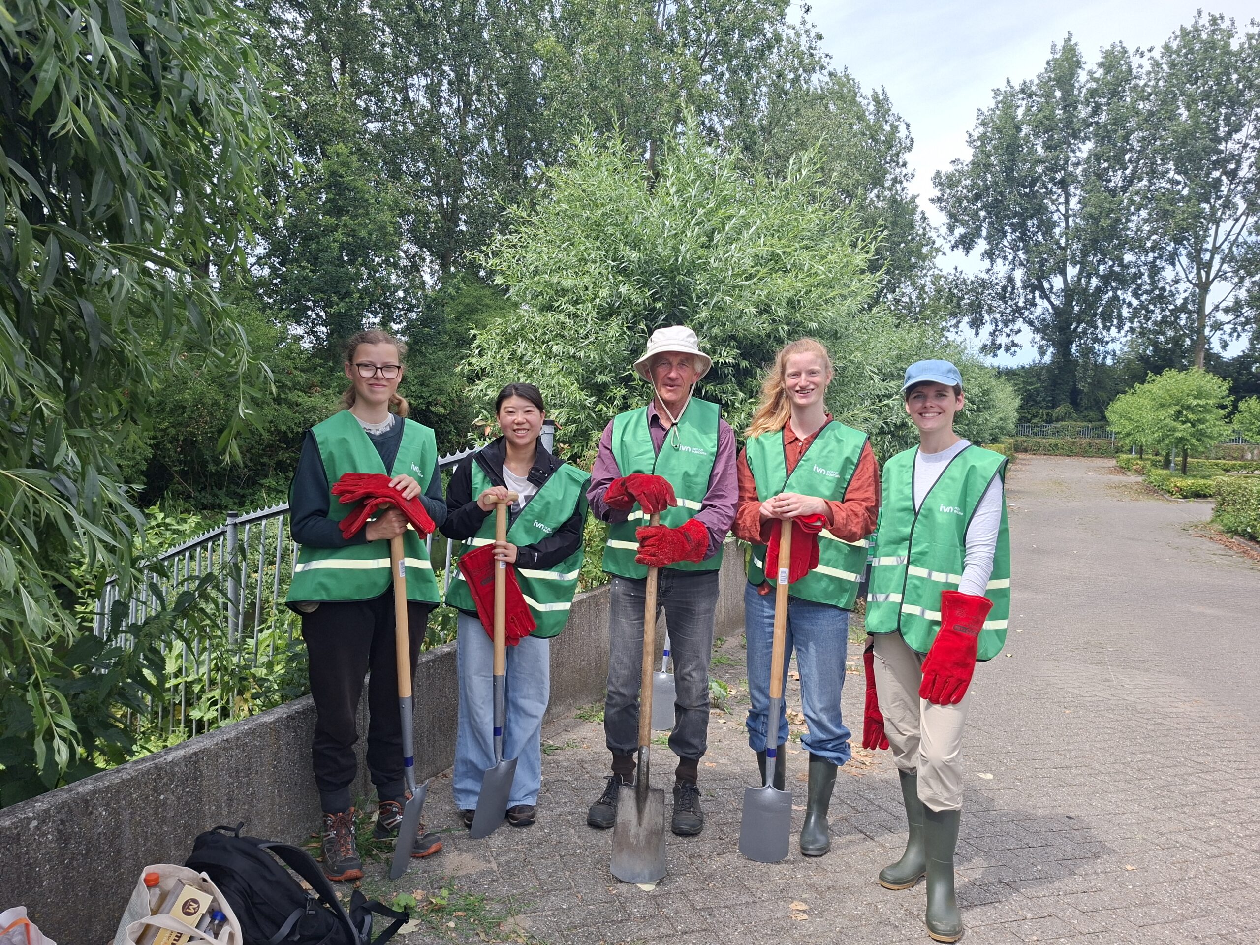 Vijf mensen in groene hesjes met scheppen en handschoenen, omringd door bomen in een park.