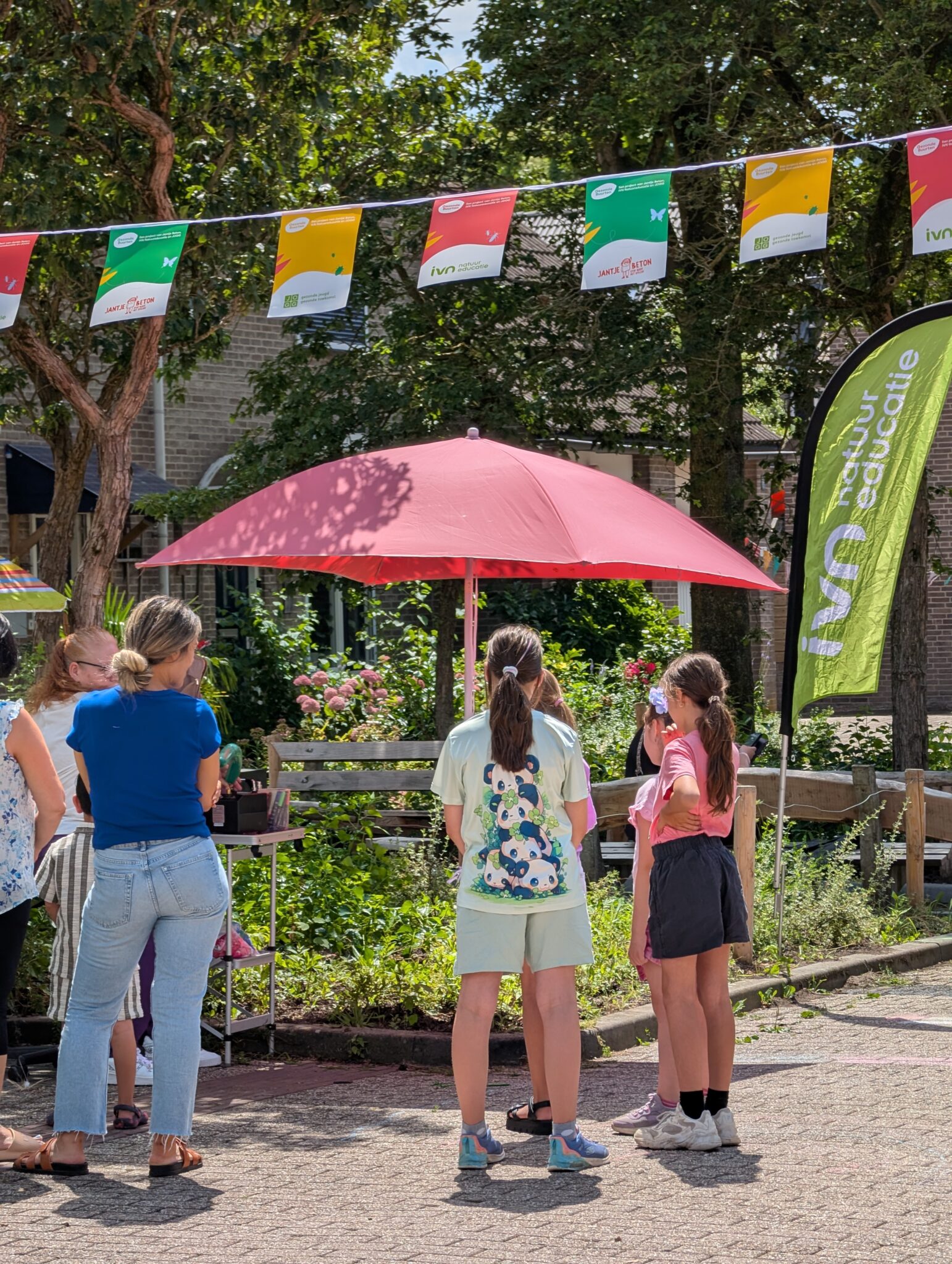 Groep mensen bij kraampje onder rode parasol, kleurrijke vlaggen erboven, buiten op zonnige dag.